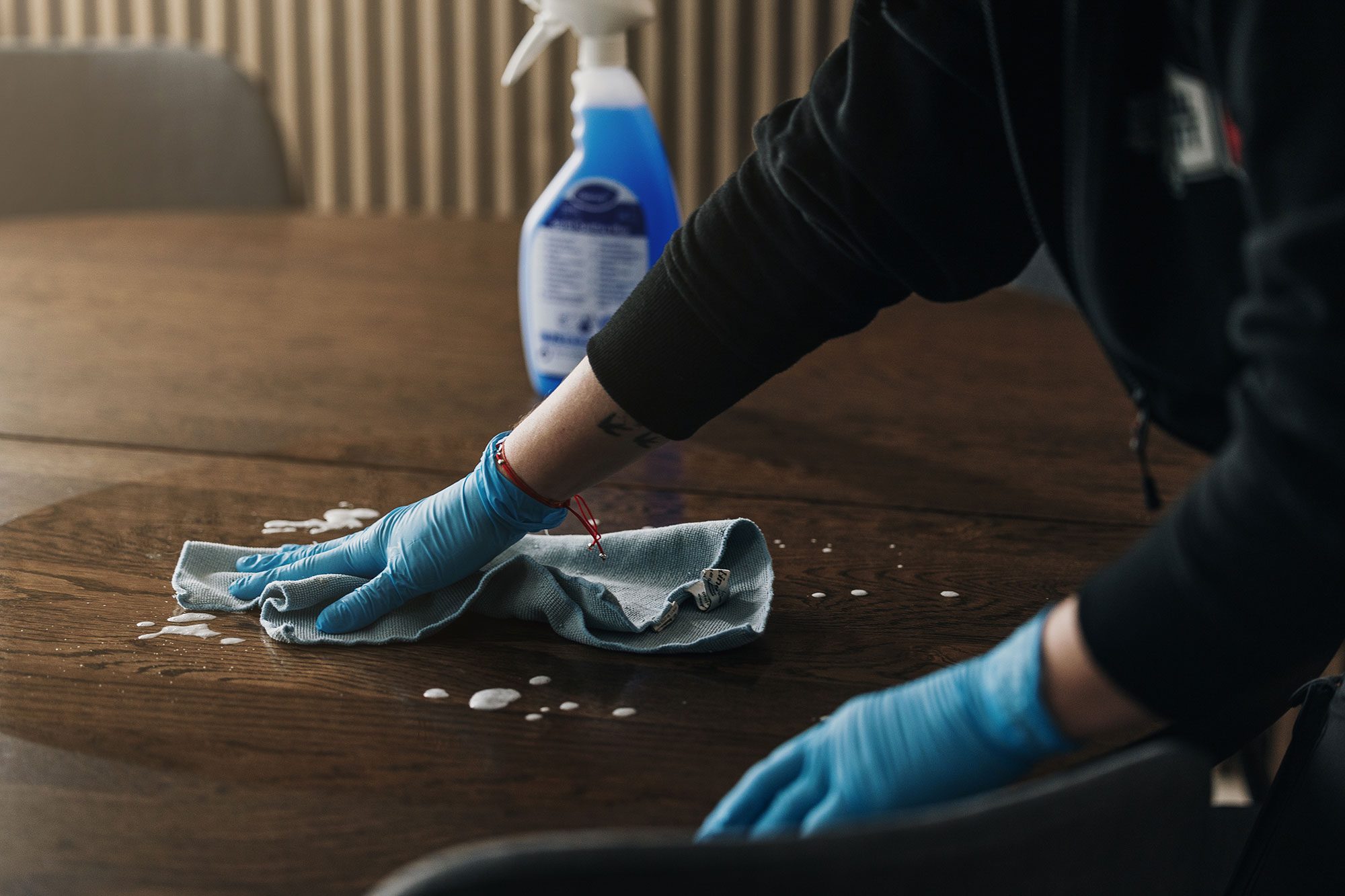 En person med blå handskar torkar av ett träbord med en trasa och rengör flytande rengöringsmedel. En sprayflaska med rengöringslösning står på bordet intill.